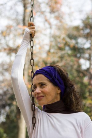 Foto de Good looking mature woman sitting on swing holding chain with one hand up, blue wrap over head and serene look. Middle aged pretty lady smiling on playground. Girl power, young at heart concepts - Imagen libre de derechos