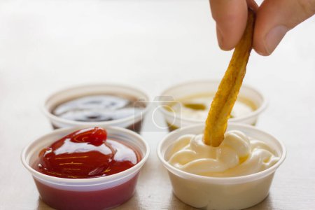 Photo for Close up on french fries dipping in mayonnaise with fingers next to plastic dips of ketchup barbecue and mustard sauce - Royalty Free Image