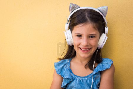 Téléchargez les photos : Sweet little girl sitting with cat ears headphones on. Cheerful kid looking at camera over yellow wall background. Pop star wannabe, fun, joy, technology, happy child concepts - en image libre de droit