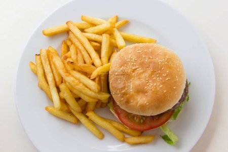 Photo for Top view of tasty beef burger with french fries on the side. Hamburger with tomato, lettuce and potatoes on white plate from above. Poor nutritional meal concept - Royalty Free Image