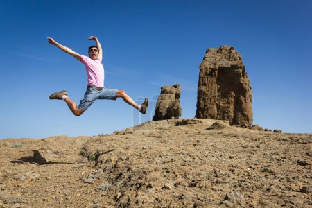 Foto de Emocionado turista saltando con las manos en alto y las piernas abiertas por el parque natural Roque Nublo, Gran Canaria. Joven alegre divirtiéndose por el punto de referencia popular. Libertad, paisaje árido, emocionados conceptos aventureros - Imagen libre de derechos