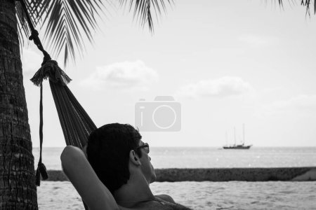 Téléchargez les photos : Young man with sunglasses resting on a beach hammock while looking at a sailboat on the background in the island of Koh Phangan, Thailand. Relax, chillout summer vacation. Black and white photography - en image libre de droit