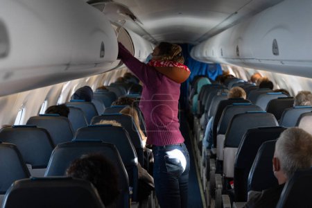 Téléchargez les photos : Femme avec coussin de cou debout sur l'allée de l'avion plaçant une valise à l'intérieur du compartiment supérieur. Prise en charge des passagers bagages à main dans l'avion - en image libre de droit