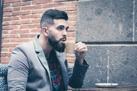 Photo for Handsome man sitting on cafe terrace sipping hot cup of coffee on street. Young hipster male model on grey blazer drinks cappuccino in the city - Royalty Free Image