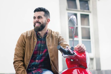 Photo for Happy young man sitting on brand new red motorcycle wearing brown leather jacket in the city. Hipster with full beard smiles on Italian classic retro scooter over white wall background - Royalty Free Image