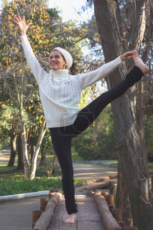 Téléchargez les photos : Smiling middle aged woman in yoga standing balance Parsva Utthita Hasta Padangusthasana. Female mature yogi on wooden path with extended hand to big toe pose. Fall season, sport activity in the park - en image libre de droit