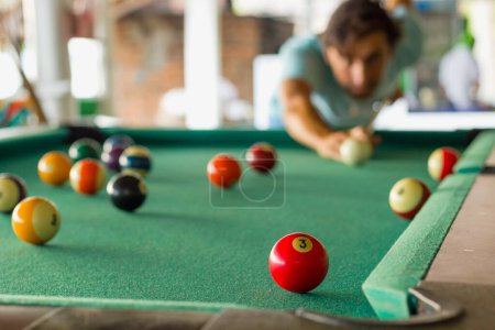 Foto de Young man playing pool with a cue aiming to strike the number three red solid ball. Billiards, snooker game concept - Imagen libre de derechos