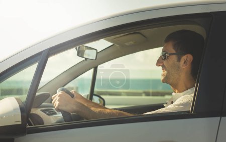 Téléchargez les photos : Happy man driving white car on sunny day by the coast with no seatbealt on. Smiling young driver with sunglasses and white shirt. Road trip, auto rental, insurance, traffic violation concepts - en image libre de droit