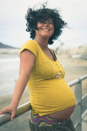 Foto de Alegre madre embarazada en camisa amarilla sosteniendo en barandilla. Jovencita sonriente esperando un bebé en un paseo por la playa en Las Palmas, España. Salud de la mujer, bienestar, conceptos de fertilidad - Imagen libre de derechos