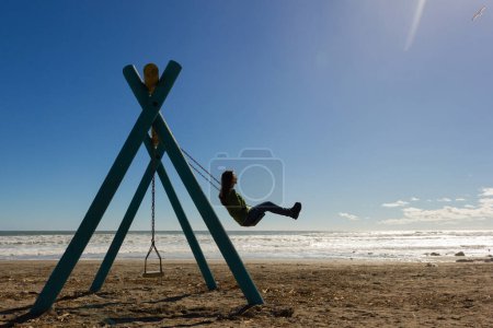 Foto de Young woman swinging on sunny winter day in La Serena beach, Chile. Child memories, empty playground, fun youth concepts - Imagen libre de derechos