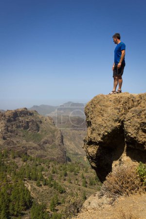 Téléchargez les photos : Young man on blue shirt looking down from top of cliff edge in Roque Nublo, Gran Canaria. Hiker on rocky mountain contemplating panoramic views. Outdoor activity, adventure, explore, freedom concepts - en image libre de droit