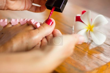 Foto de Close up on foot of a woman meticulously painting her big toenail with nail polish standing by tropical leelawadee flower. Pedicure, nails salon concept - Imagen libre de derechos