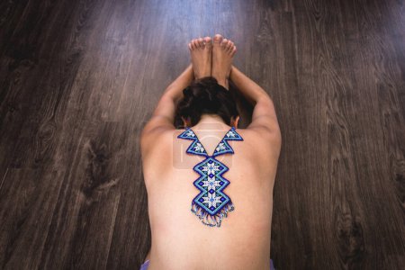 Foto de Top less woman on paschimottanasana with huichol blue necklace on naked back. Female yogi on seated forward bend on wood planks floor. Fashion design, Mexican handcraft, colorful decoration concepts - Imagen libre de derechos