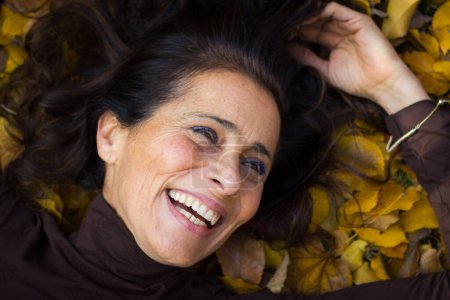 Téléchargez les photos : Lovely portrait of middle aged gorgeous woman with big smile while lying on ground full of yellow and brown leaves in the park during autumn season. Soft vintage effect - en image libre de droit