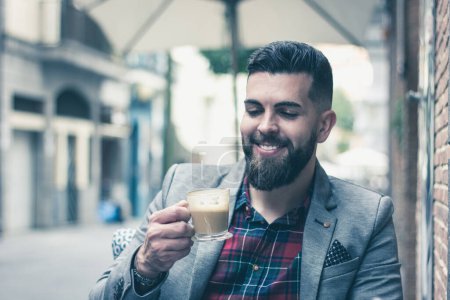 Téléchargez les photos : Young businessman loving his coffee on street terrace cafe. Smiling handsome man with grey blazer looking at cup of espresso with satisfaction expression. Delicious cappuccino, leisure time concept - en image libre de droit