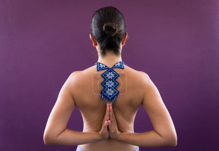 Photo for Top less woman on pashchima namaskarasana over purple wall with huichol necklace on naked back. Female yogi on reverse prayer pose with traditional handcraft from Mexico. Penguin pose, fashion concept - Royalty Free Image