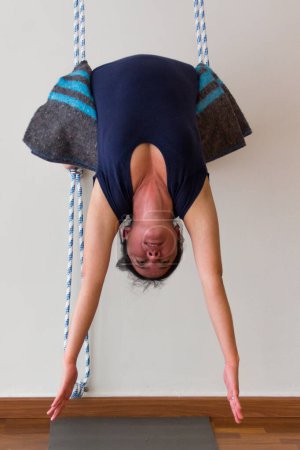Foto de Yoga teacher on upside down posture resting on blanket and grabbed by ropes on white wall background. Young female yogi practices backbend advanced asanas in studio - Imagen libre de derechos