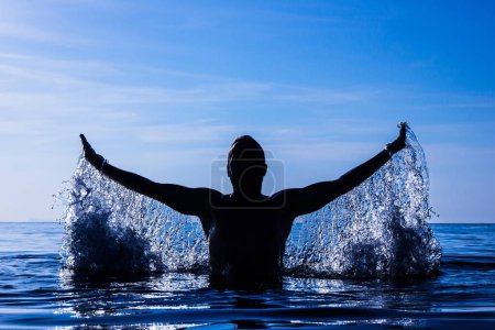 Téléchargez les photos : Mouvement de gel de la silhouette du jeune homme éclaboussant l'eau à bras ouverts sur l'eau au coucher du soleil mystique dans l'île de Koh Phangan, Thaïlande. Filtre bleu foncé. Victoire, triomphe, concepts gagnants - en image libre de droit