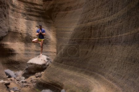 Téléchargez les photos : Young man on vrikshasana asana standing on rock in flowing shapes wall canyon. Hands on namaste. Yogini practices yoga on arid environment. Tree pose. Outdoor activity. Hope concept - en image libre de droit