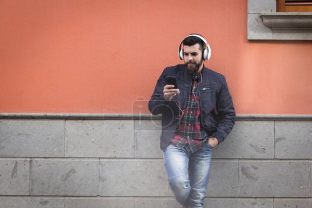 Photo for Modern looking young man with headphones leaning on brown and gray wall. Hipster male model using smart phone to select music from playlist - Royalty Free Image
