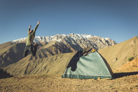 Photo for Big jump of energetic young man next to igloo camping tent with magnificent views of Andes snowed mountain range on the background under blue sky in Elqui Valley, Chile. Explore, hiking concepts - Royalty Free Image