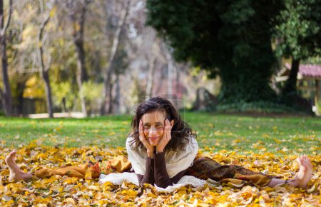Téléchargez les photos : Flexible middle aged woman with open legs and hands on her face smiles sitting on brown and yellow leaves in the park. Pretty mature lady posing in autumn season outdoors. Healthy lifestyle concept - en image libre de droit