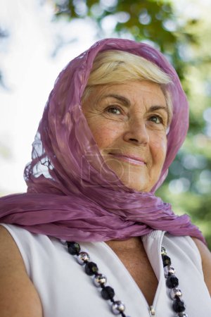 Foto de Portrait of mature lady covering her head with hijab style shawl in the park. Young looking woman with purple veil over head, Muslim look. Female model with charming serene expression and soft skin - Imagen libre de derechos