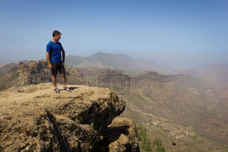 Photo for Tourist on mountain edge facing great landscape views. Young man on blue t shirt standing on top of rock on sunny day in Roque Nublo. Adventure, explore, freedom, visionary, solitude concepts - Royalty Free Image