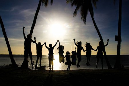 Foto de Siluetas de un grupo de niños y jóvenes saltando en el aire mientras se toman de la mano al atardecer con el mar al fondo en la pequeña isla de Malapascua, Filipinas. - Imagen libre de derechos