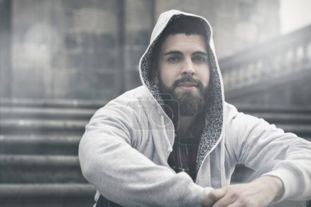 Photo for Young male model with full beard covers head with grey hoodie sitting on stairs in old town. Hip hop, rapper style concept. Sun flare effect - Royalty Free Image