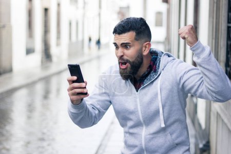 Téléchargez les photos : Enthusiastic young man looking at cellphone with victorious expression in the city. Overexcited hipster with grey hoodie holding mobile device and fist up. Online bet winner concept - en image libre de droit