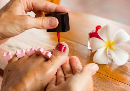 Foto de Close up on foot of a woman meticulously painting her big toenail with nail polish standing by tropical leelawadee flower. Pedicure, nails salon concept - Imagen libre de derechos