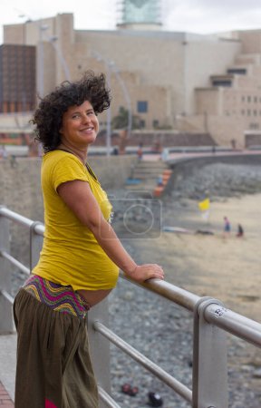 Foto de Feliz mujer embarazada con camisa amarilla sosteniendo la barandilla. Dama sonriente esperando bebé en paseo por la playa en Las Palmas, España - Imagen libre de derechos