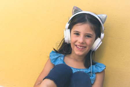 Téléchargez les photos : Douce petite fille sourit avec des écouteurs chat. Portrait d'enfant joyeux regardant la caméra sur fond de mur jaune. Pop star aspirant, amusant, joie, technologie, concepts enfants heureux - en image libre de droit
