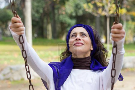 Téléchargez les photos : Modern good looking mature woman holding to chains while swinging in the playground wearing purple shawl on head. Middle aged pretty lady remembering child memories in the park. Young at heart concept - en image libre de droit