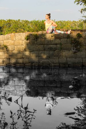 Photo for Yogi woman in marichyasana c pose on top of rocks wall in color with reflection on black and white on river by Srirangapatna, Karnataka, India. Lady practices yoga in nature, healthy lifestyle concept - Royalty Free Image