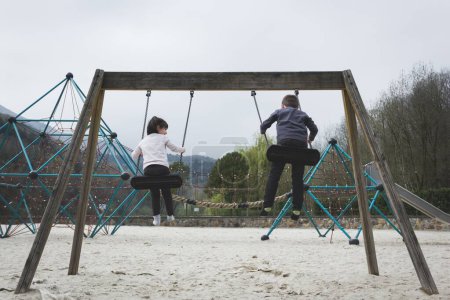 Téléchargez les photos : Quelques petits enfants s'amusent à se balancer à l'aire de jeux par temps nuageux à Bilbao. Deux enfants jouent dans le parc. Tonalités de filtre à froid appliquées - en image libre de droit