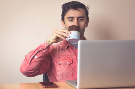 Téléchargez les photos : Young man with red shirt drinking coffee while looking at screen of laptop at office desktop. Cellphone on table. Own boss working remotely from home concept - en image libre de droit