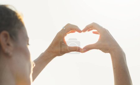 Foto de Woman with hands in heart shape on sunny day. Young lady with romantic symbol at twilight. Valentine Day, sunny, healthy lifestyle concepts - Imagen libre de derechos
