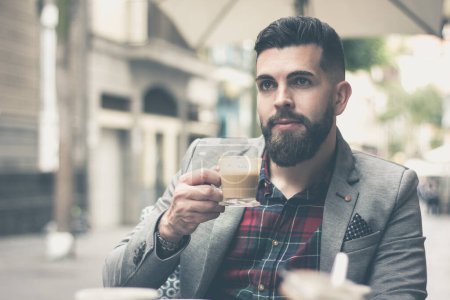 Photo for Attractive man holding cup of coffee on cafe terrace in the city. Handsome businessman with full beard and grey blazer taking a break with cappuccino. Young male model enjoying espresso on street - Royalty Free Image