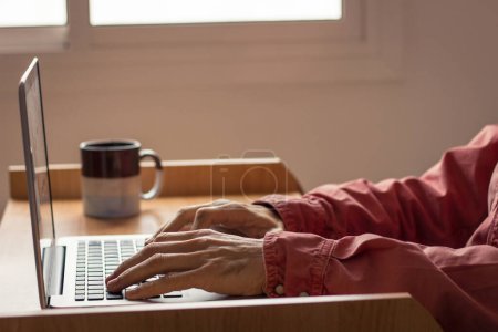 Photo for Hands of man typing on laptop with coffee mug next to him on desktop. Wearing long sleeve red shirt. Work remotely from home concept. Freelance, web designer job - Royalty Free Image