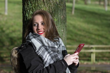Téléchargez les photos : Smiling cute girl looks over her shoulder while holding red smart phone leaning on tree trunk in the park. Beauty fashion model, autumn sales concept. Young pretty woman with cellphone - en image libre de droit