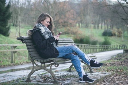 Téléchargez les photos : Pretty girl sitting on bench in park smiles at camera while holding mobile phone. Fashion trend of millennial generation on cellphone addiction - en image libre de droit