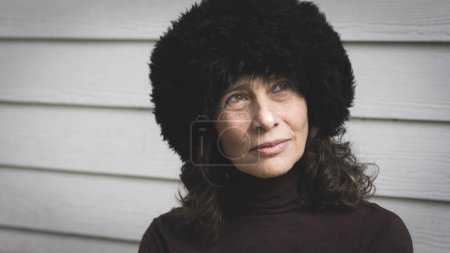 Photo for Mature good looking woman with black Russian hat and thoughtful gaze on white wood planks background. Portrait of middle aged beautiful lady on winter accessories. Cold winter weather concept - Royalty Free Image