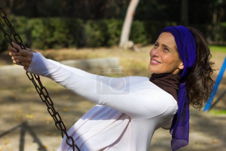 Foto de Happy beautiful woman with purple shawl on head swinging in the playground on sunny day. Care free charming mature lady remembering the old days, freedom, young spirit concepts - Imagen libre de derechos