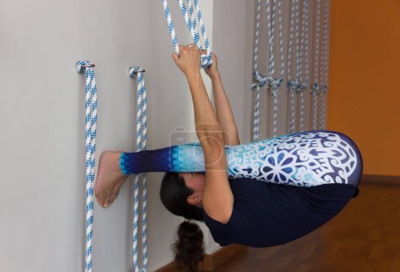 Foto de Yoga teacher in blue sportswear practices paschimottanasana inverse pose grabbing to ropes on wall. Iyengar practitioner doing calisthenics exercise in yoga studio. Flexibility concept - Imagen libre de derechos