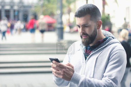 Téléchargez les photos : Full bearded man using cellphone outdoors in the city. Young hipster with gray hoodie sending text message with black mobile device on street. Communication, wireless internet concept - en image libre de droit