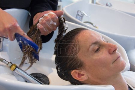 Foto de Mujer joven que se lava la cabeza y peluquería tratando de desenredar el cabello con peine. Cliente femenino en el salón de belleza - Imagen libre de derechos