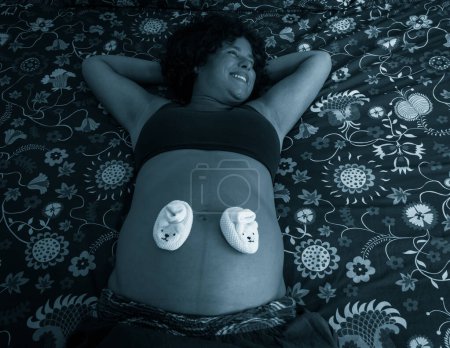 Téléchargez les photos : Heureuse femme enceinte couchée sur un lit de housse de couette design abstrait avec des chaussures de bébé sur le dessus du ventre. Jeune femme souriante se reposant dans la chambre. Effet bleu appliqué - en image libre de droit