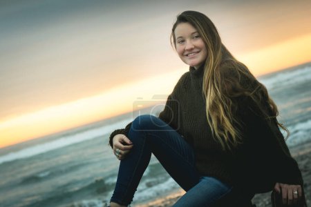 Téléchargez les photos : Fille blonde souriante pose assis sur des piliers en bois sur la plage au coucher du soleil jour d'hiver à La Serena, Chili. Jeune femme au bord de la mer au crépuscule. Horizons inclinés - en image libre de droit